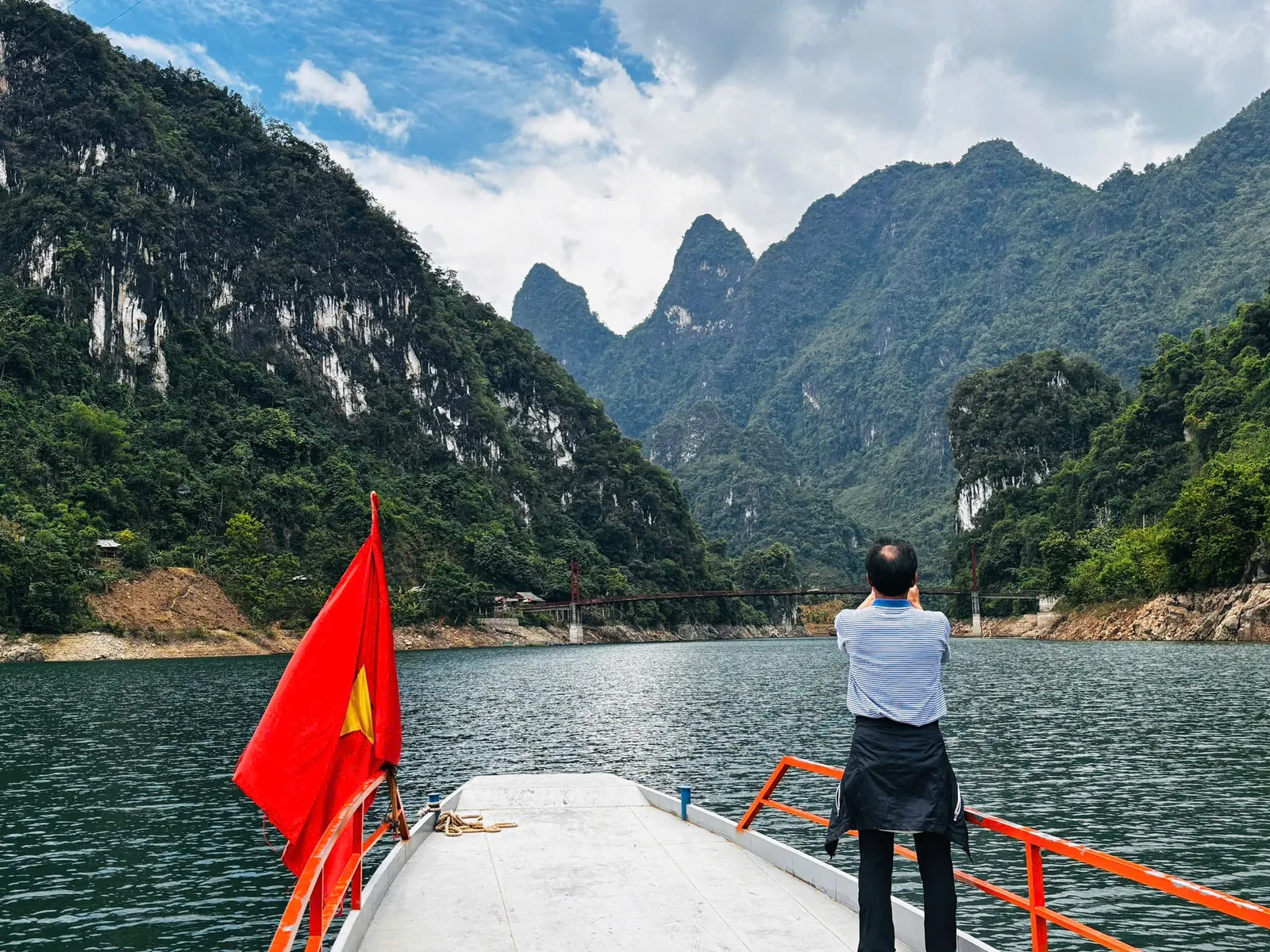 Du khách chụp ảnh từ mũi thuyền cắm cờ đỏ sao vàng trên hồ giữa núi đá vôi Du khách chụp ảnh từ mũi thuyền cắm cờ đỏ sao vàng trên hồ giữa núi đá vôi